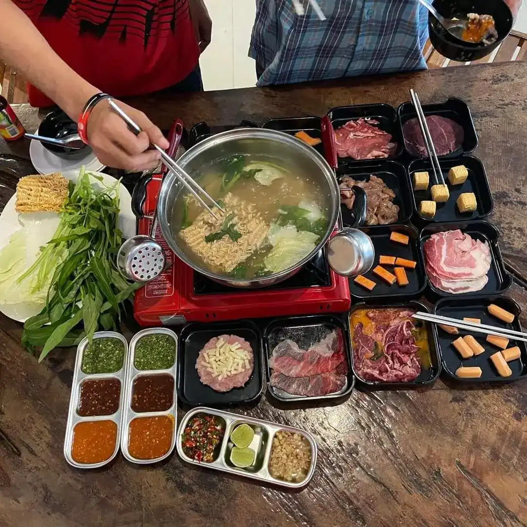 A shabu set seen from above: a pot of broth with noodles on a table burner, trays of sliced meat, tofu and vegetables, and three dipping sauces