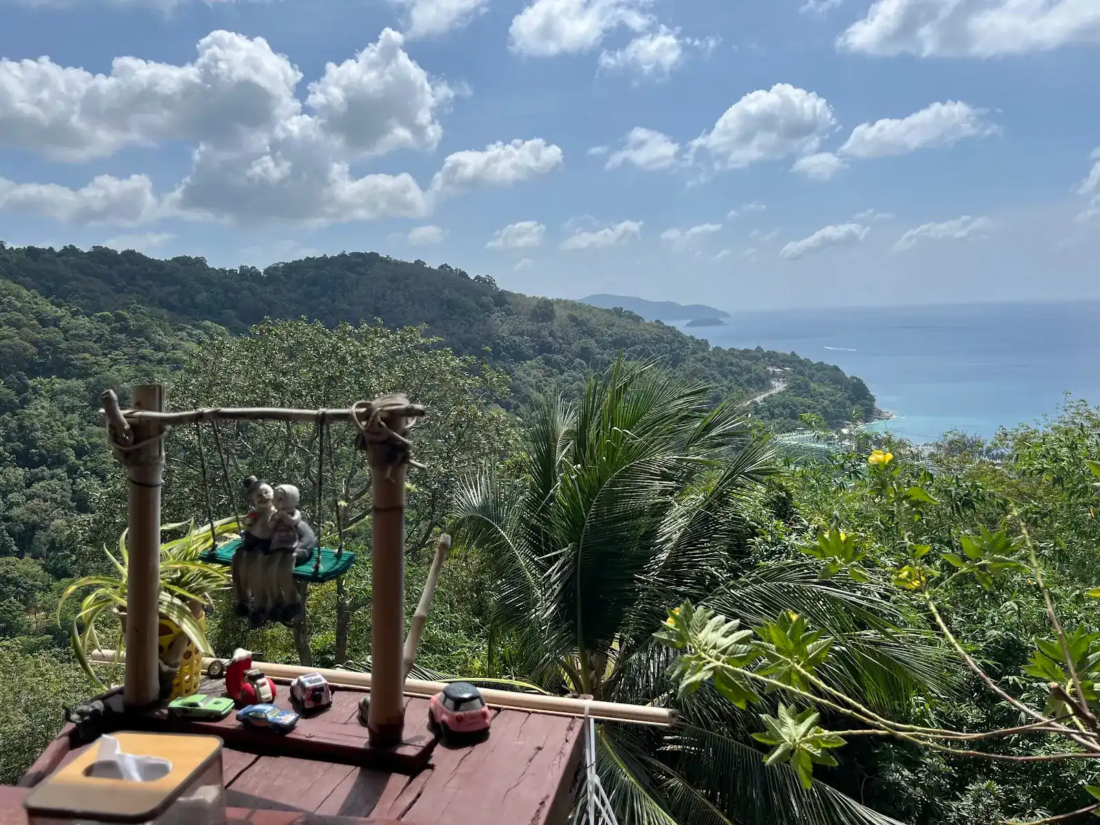The view from the terrace: jungle hills running down to the coast road and the blue sea, with a little grandma-and-grandpa figurine on a swing