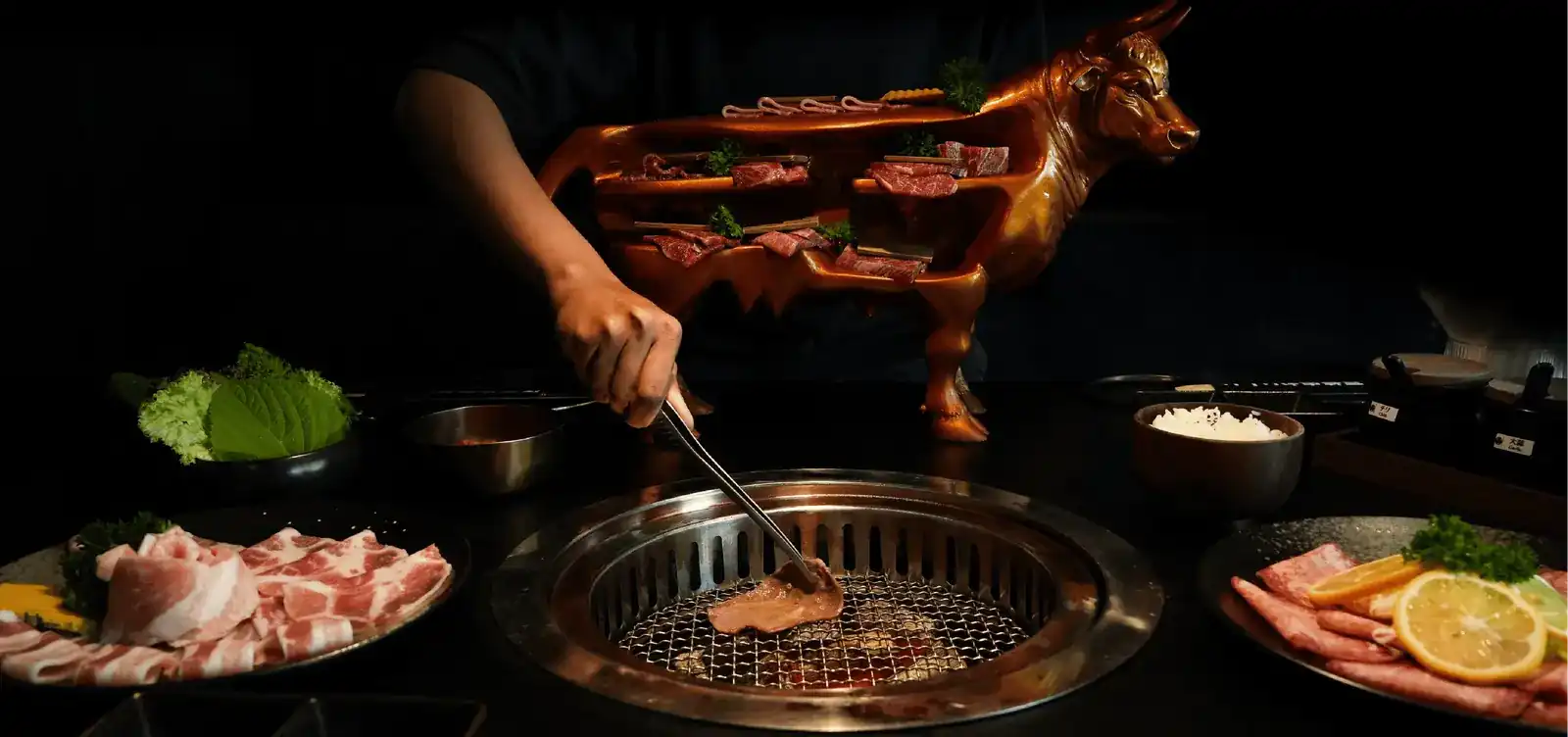 A hand turns a slice of beef on a round charcoal grill; behind it a bronze bull carries shelves of raw cuts