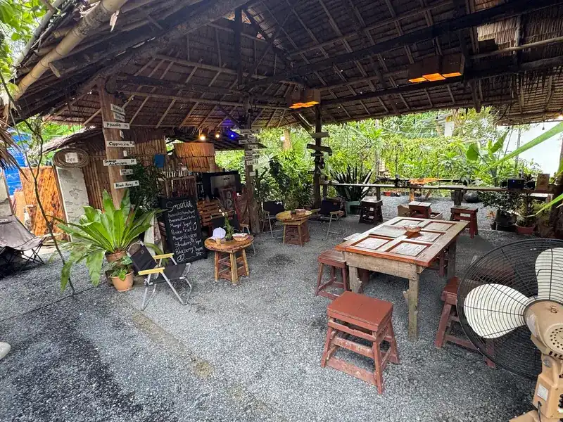 Open-air seating under a thatched roof: wooden tables and stools on a gravel floor among plants
