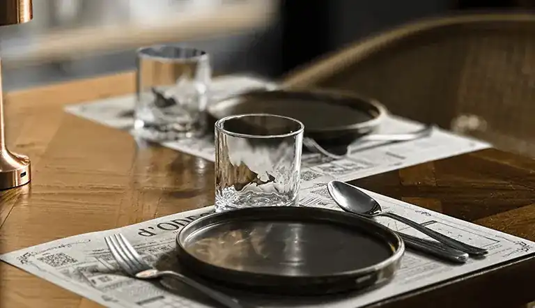 A table set with dark plates on newspaper-print placemats / โต๊ะที่จัดจานสีเข้มบนแผ่นรองจานลายหนังสือพิมพ์