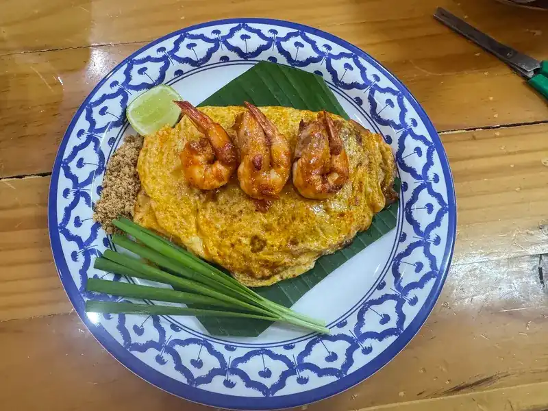 Pad thai wrapped in egg, topped with three prawns, on a blue and white plate with lime and chives