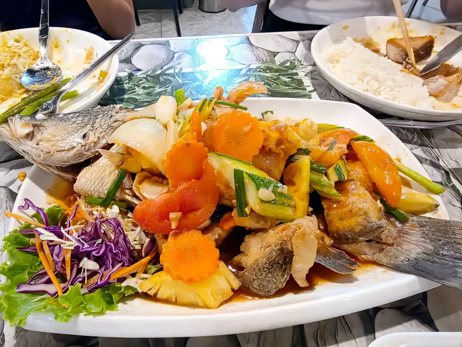 A whole fried fish topped with pineapple, tomato, cucumber, carrot and onion in a glossy sauce, on a pebble-print table