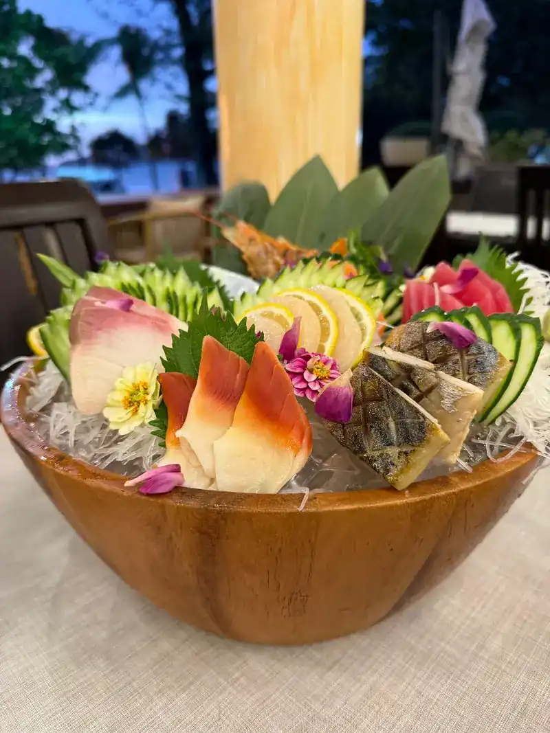 A sashimi platter on ice in a wooden bowl, with palm trees and the sea behind at dusk