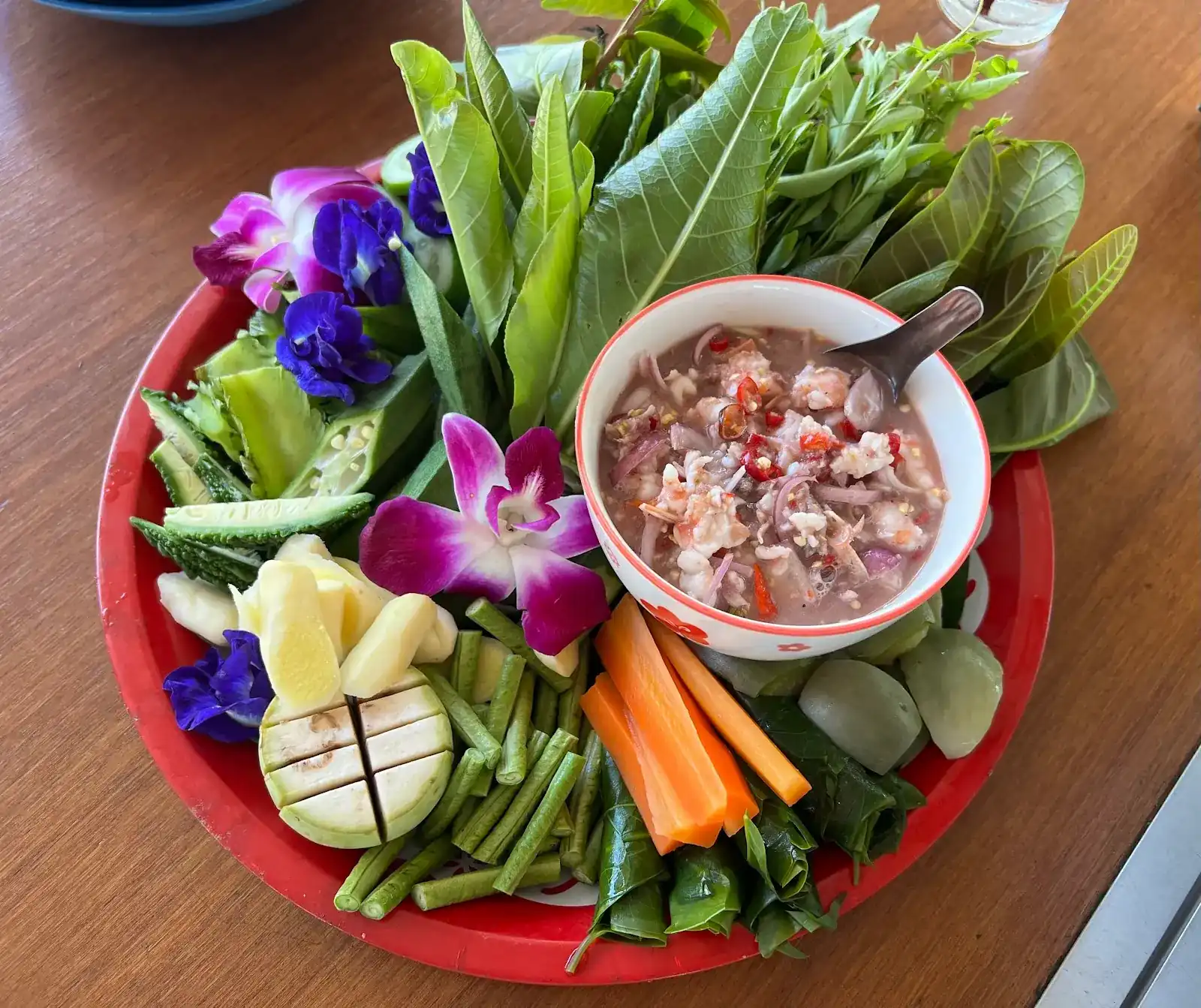 A bowl of chilli dip with prawns on a red tray, ringed by fresh leaves, long beans, carrot, cucumber, orchids and butterfly pea flowers