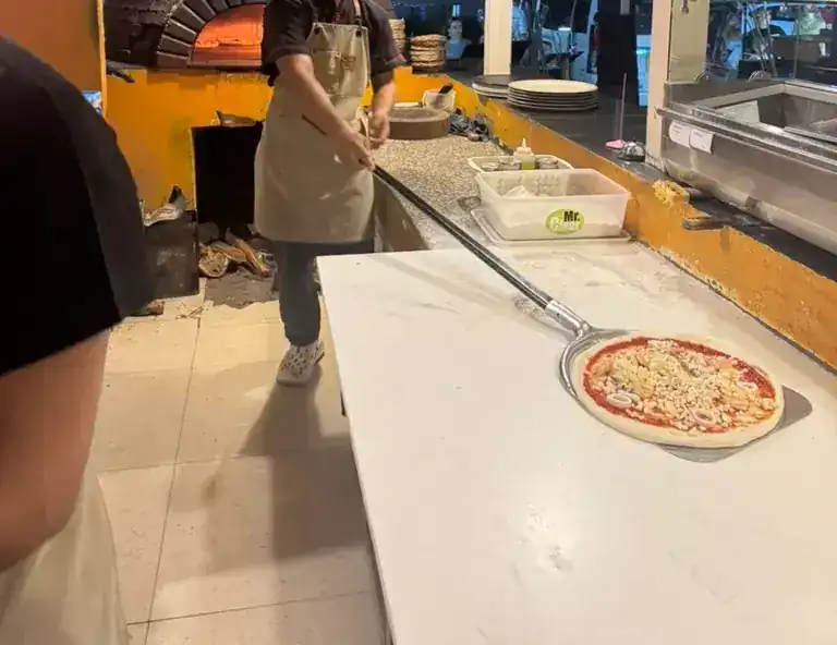 A pizza on the peel at the counter, with the brick oven glowing behind