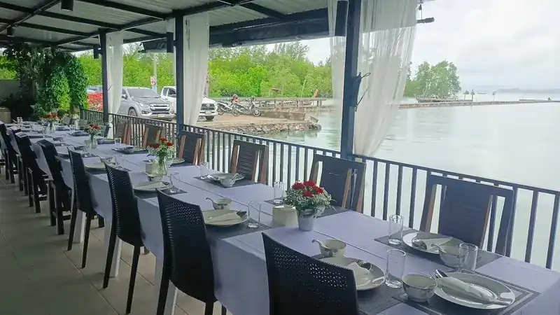 A long table set in lilac cloth along the rail of the covered deck, calm water and mangroves beyond