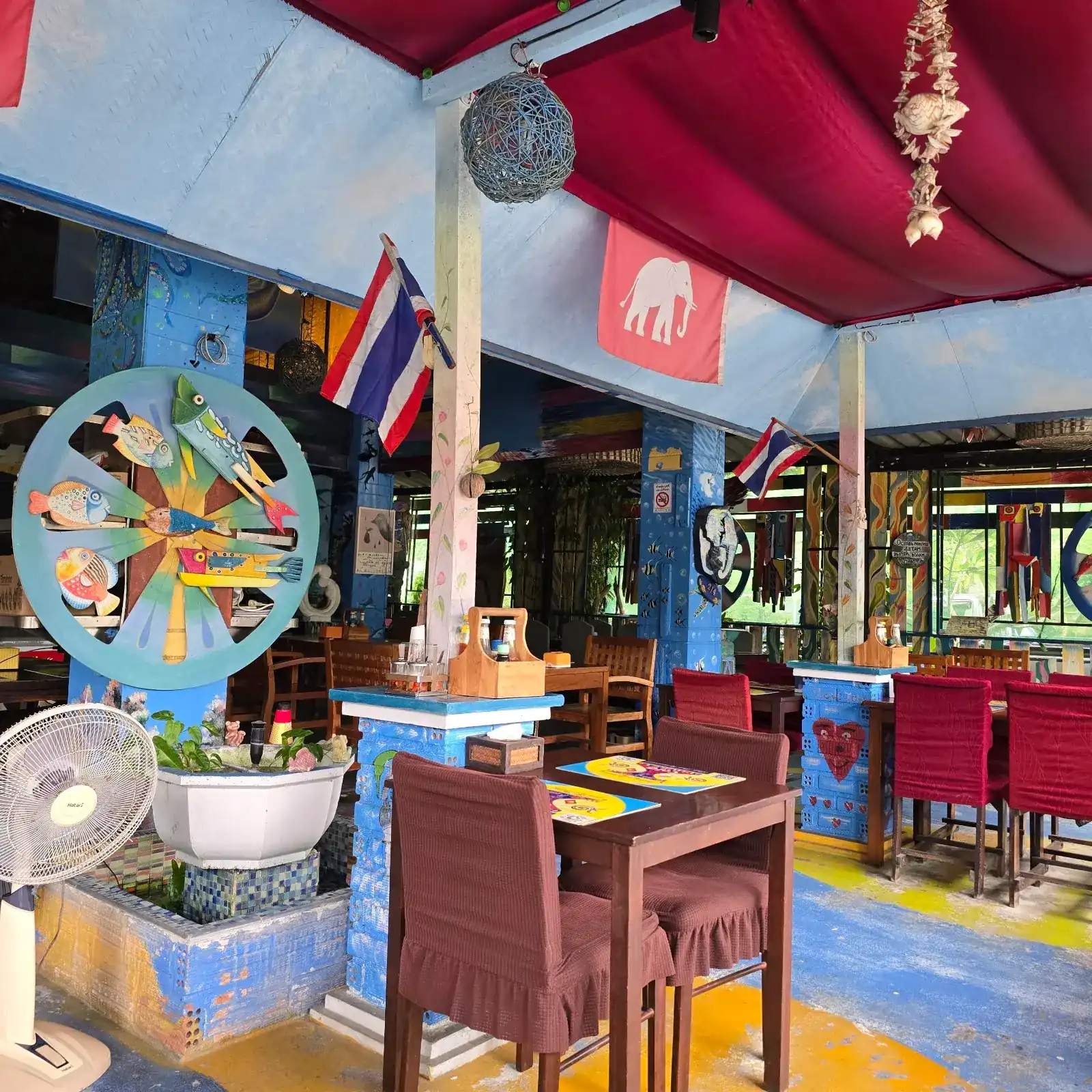 Tables under a red canopy at Thai Thai Restaurant, with a white-elephant flag, Thai flags and a wheel of painted wooden fish