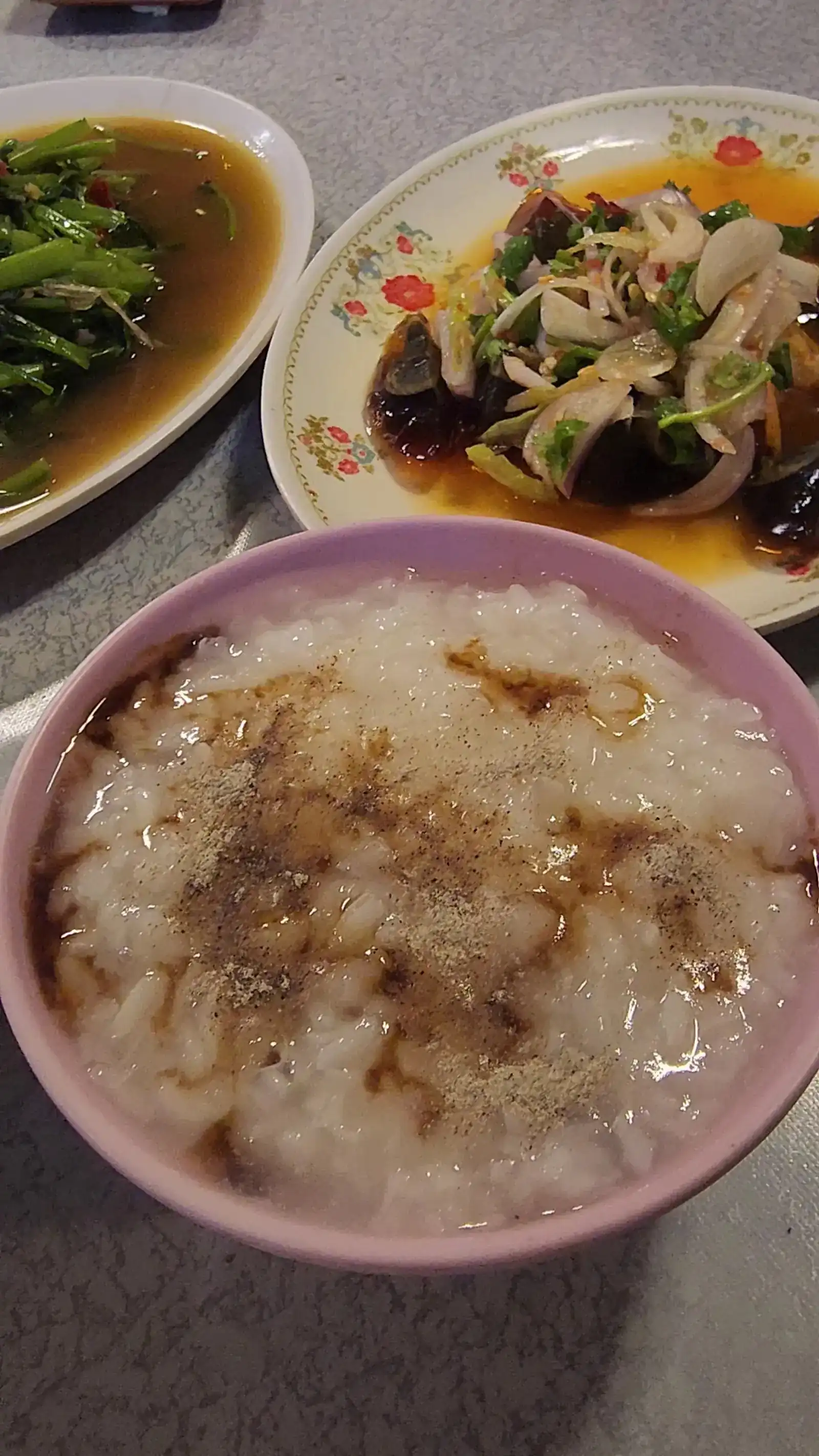 A pink bowl of plain rice soup sprinkled with pepper, with stir-fried water spinach and a preserved egg salad behind it