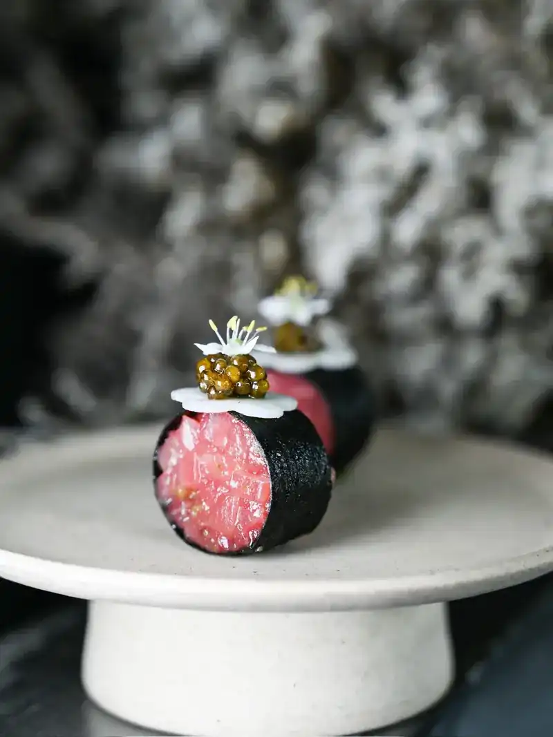 A dark-wrapped roll of raw fish topped with caviar and a white flower, on a stone stand