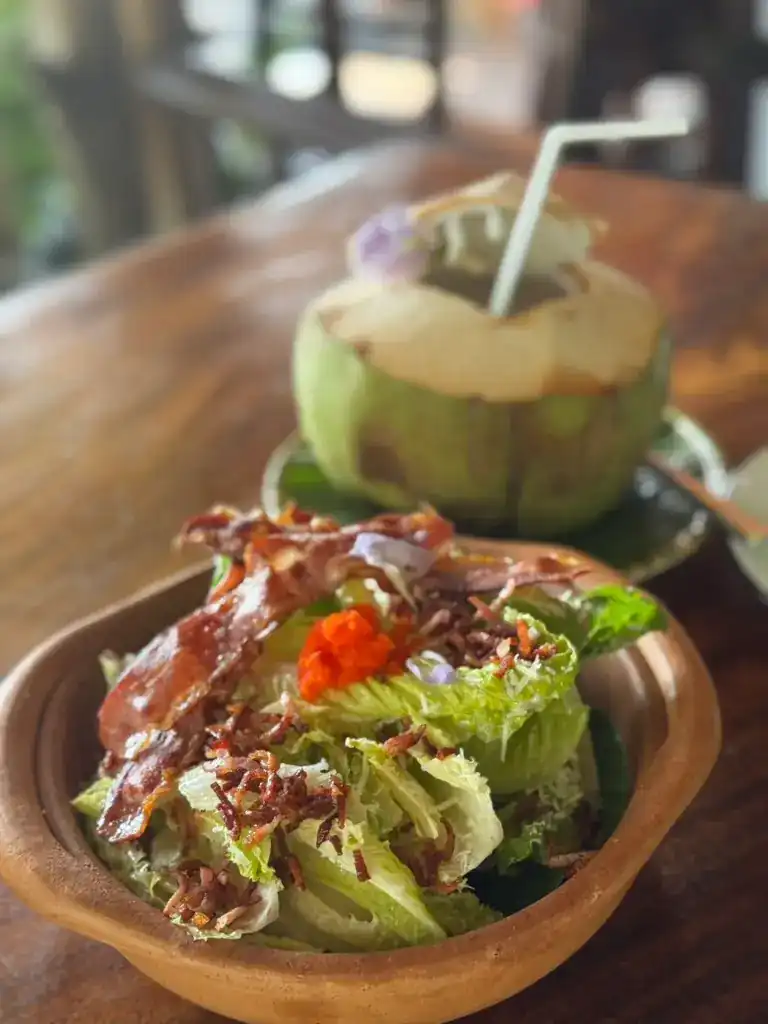 A salad with crispy bacon in a clay bowl, a fresh coconut behind