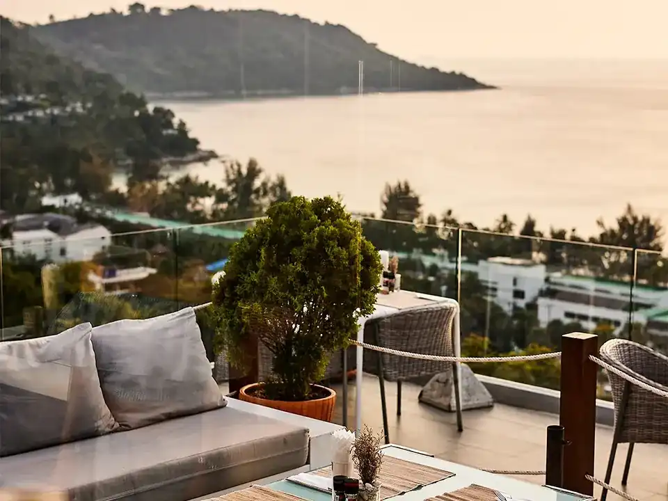 Sofa and tables on the terrace behind a glass rail, the bay and headland of Kata Noi in evening light
