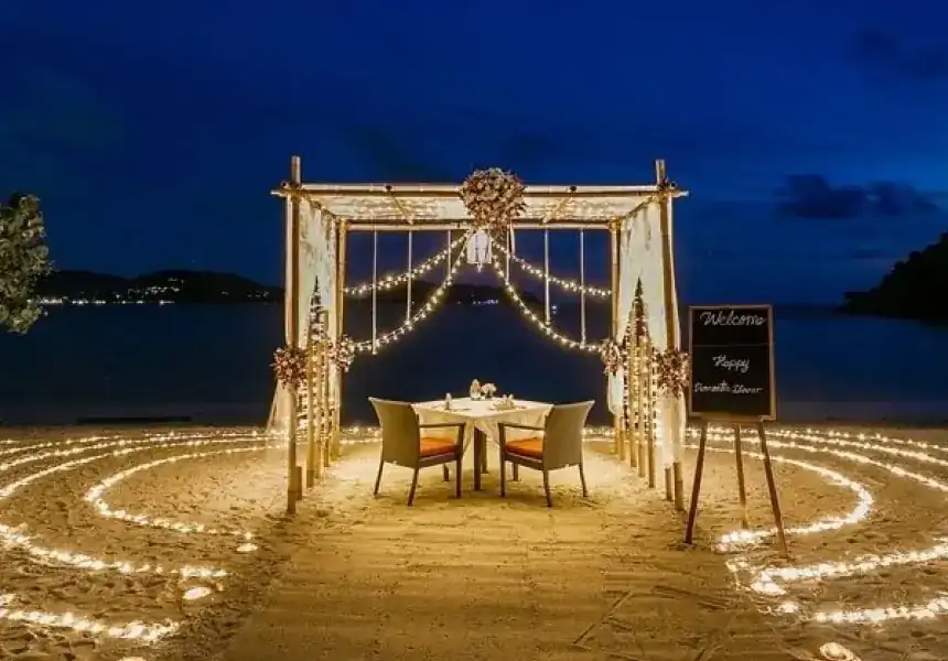 A table for two under a bamboo cabana on the sand at night, ringed by circles of fairy lights