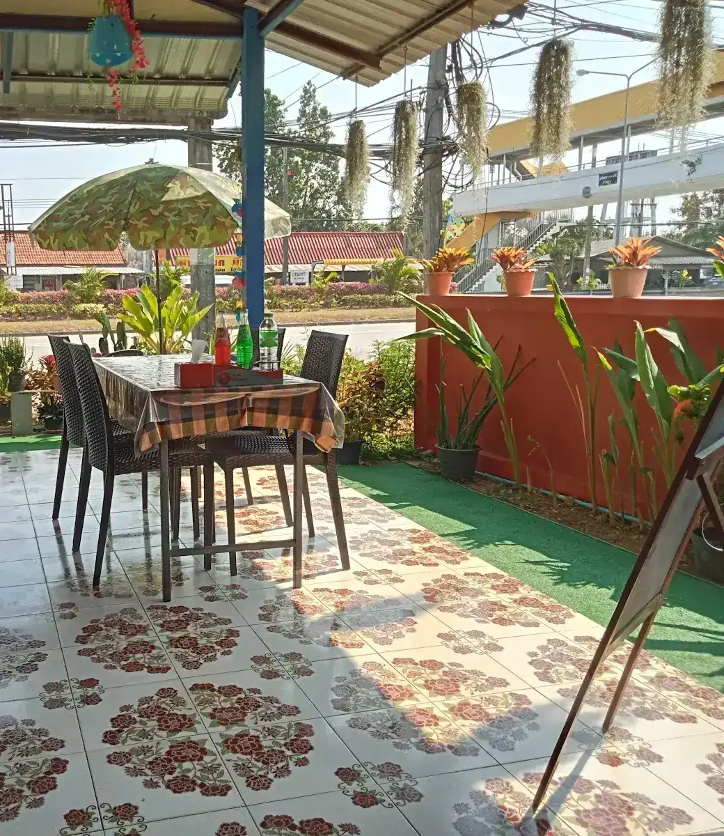 The covered terrace by the road: rose-patterned floor tiles, a table under a parasol, sky-blue posts / ลานใต้หลังคาริมถนน พื้นกระเบื้องลายดอกกุหลาบ โต๊ะใต้ร่ม และเสาสีฟ้า