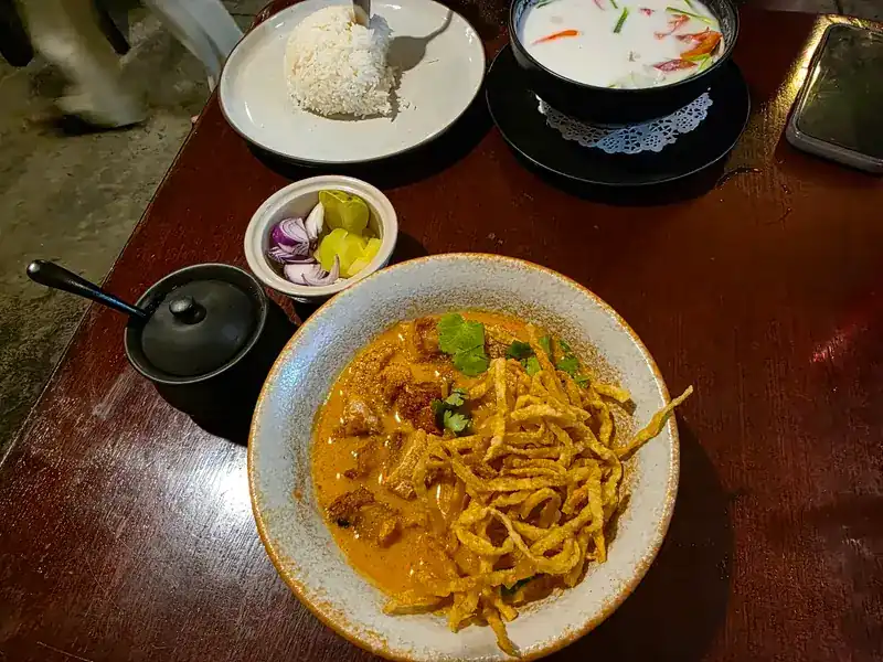 Khao soi topped with crispy noodles and coriander, with pickled greens, shallots, rice and a coconut soup on a wooden table