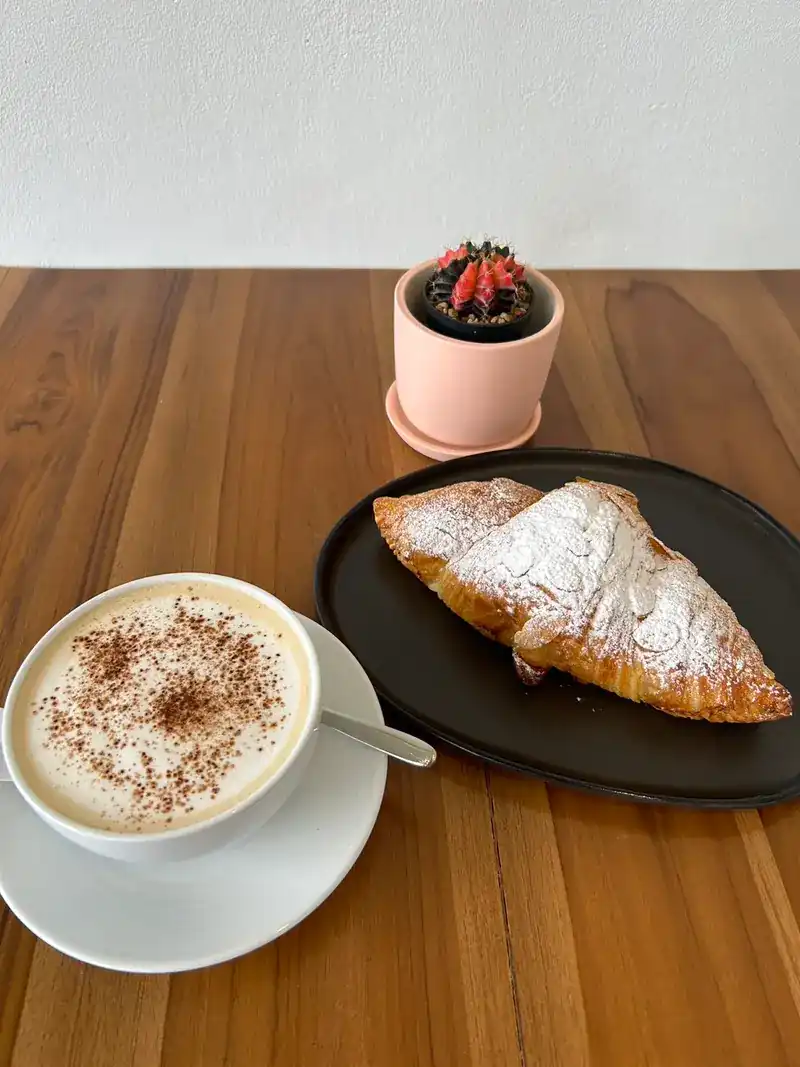 A croissant dusted with sugar on a black plate, next to a cappuccino