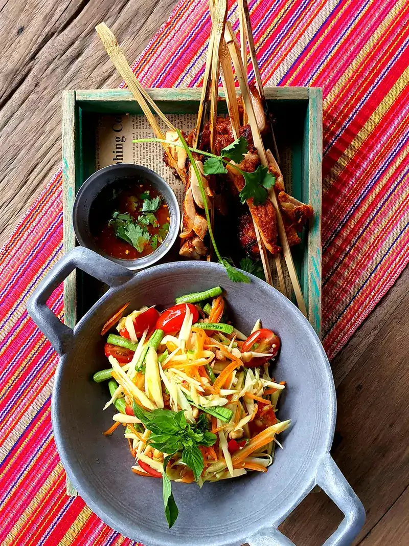 Chicken grilled in split bamboo beside a shredded salad in a grey pan, on a striped woven cloth