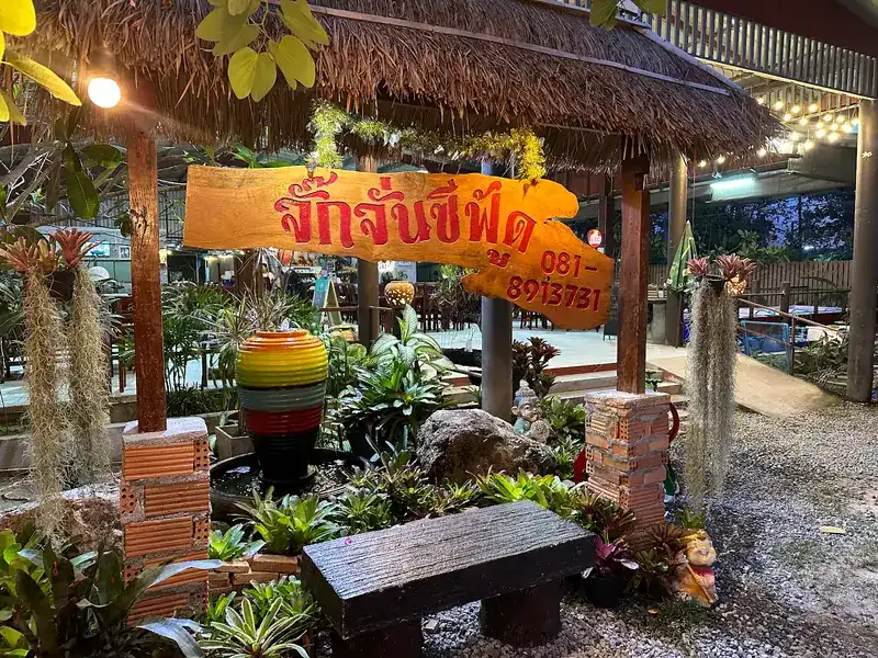 A carved wooden sign with the restaurant’s name under a thatched roof, in a lit garden in the evening