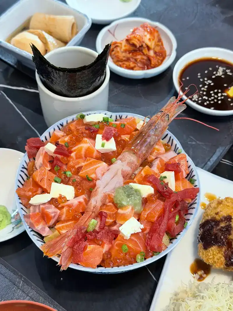 A bowl heaped with salmon and tuna, topped with a whole prawn, kimchi and nori beside it