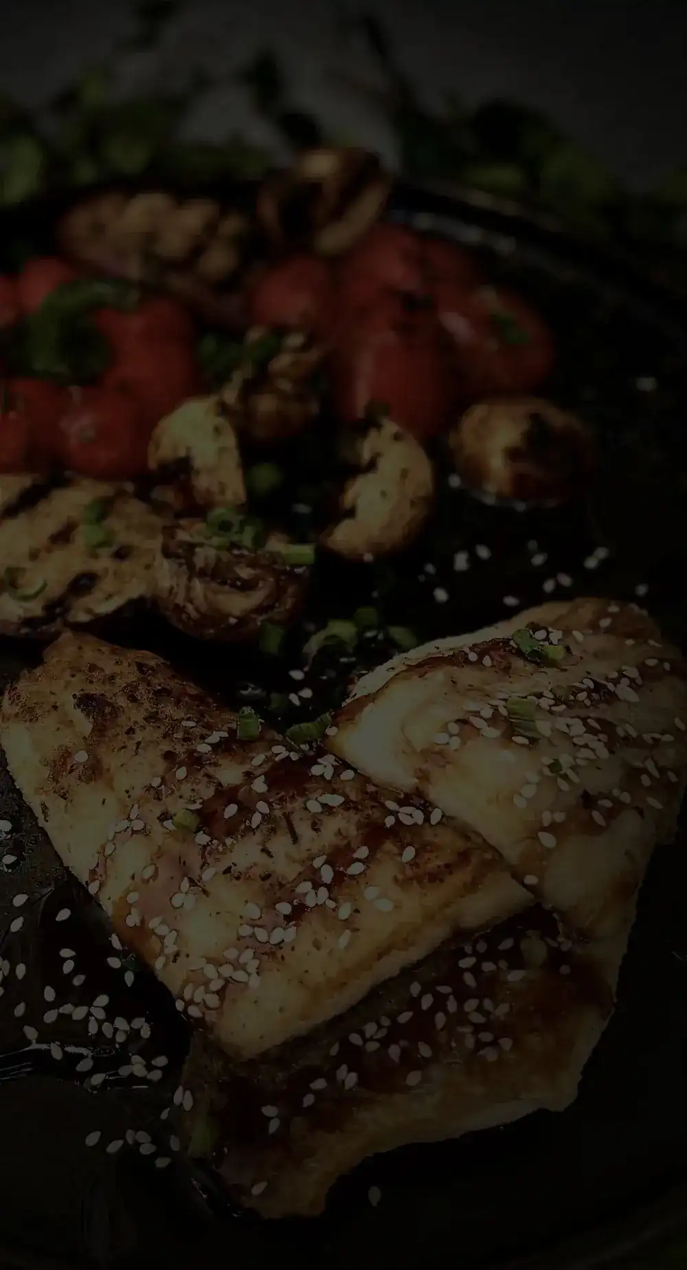 Grilled fillets scattered with sesame, with roasted tomatoes and mushrooms, on a dark plate