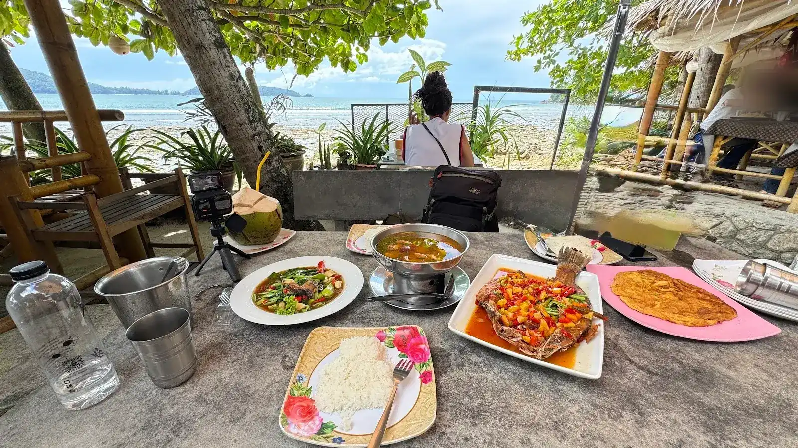 A table at the edge of the beach with a whole fish in chilli sauce, soup on a burner, stir-fried greens, an omelette, rice and a coconut, the sea behind