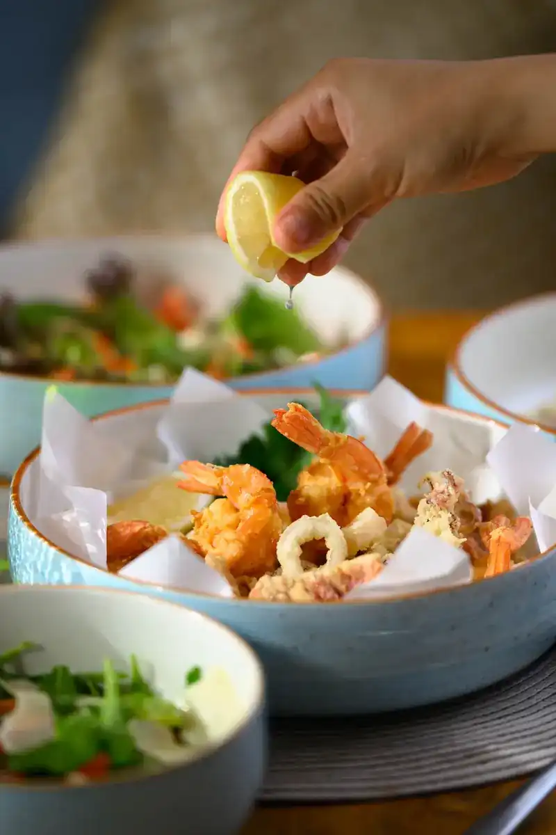 A hand squeezing lemon over fried prawns and calamari in a blue bowl