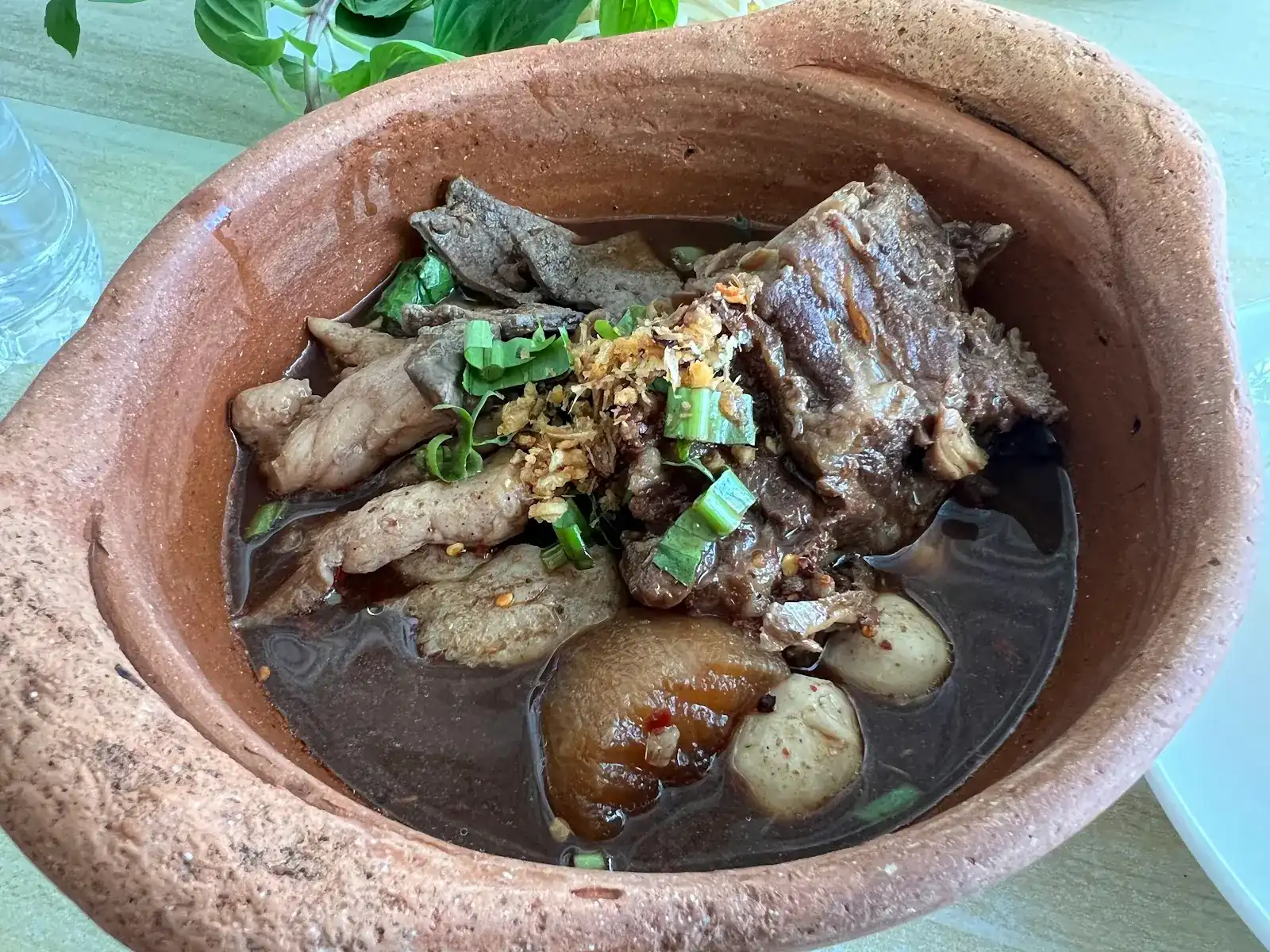 Dark boat-noodle broth in a clay pot with pork, meatballs, spring onion and fried garlic