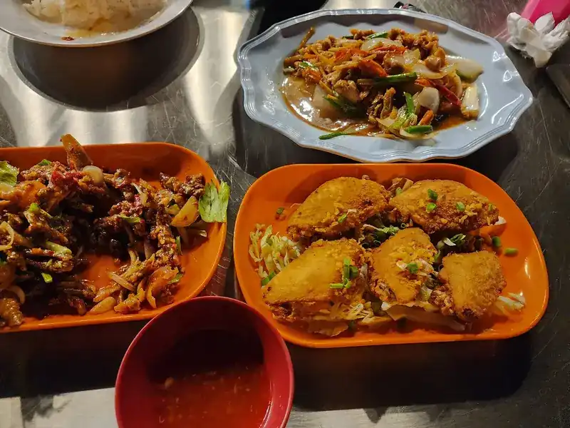 Three plates on a steel table, a spicy salad, battered fried pieces and a stir-fry, with a bowl of chilli sauce