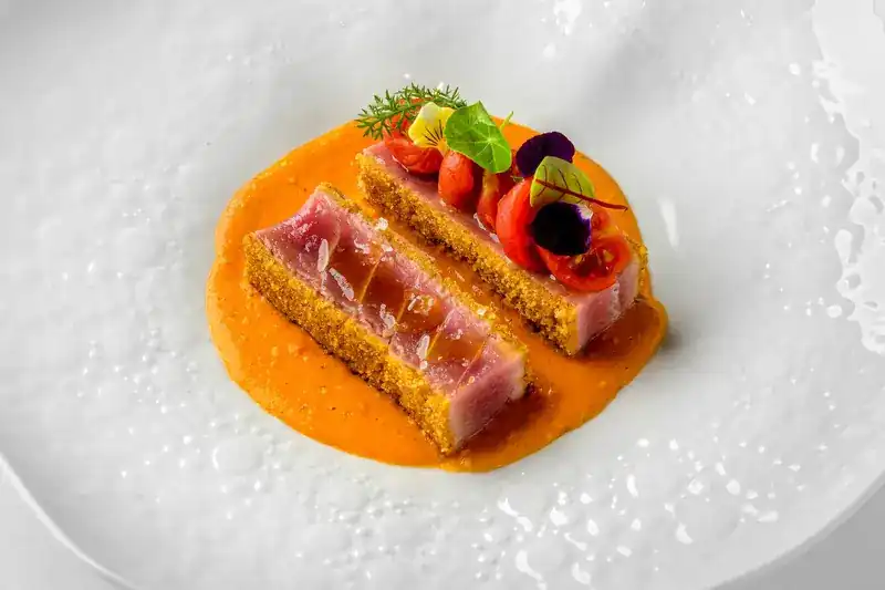 Two crusted slices of seared tuna on an orange sauce with flowers and herbs