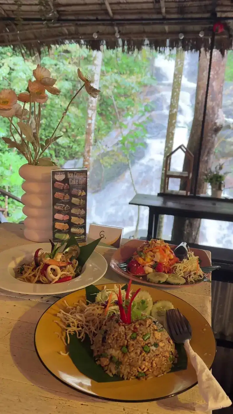 Three plates of Thai food, one a fried noodle dish, on a table under the thatch roof with the waterfall behind