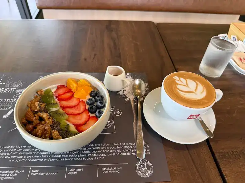 A bowl with strawberries, kiwi, blueberries and nuts, next to a cappuccino