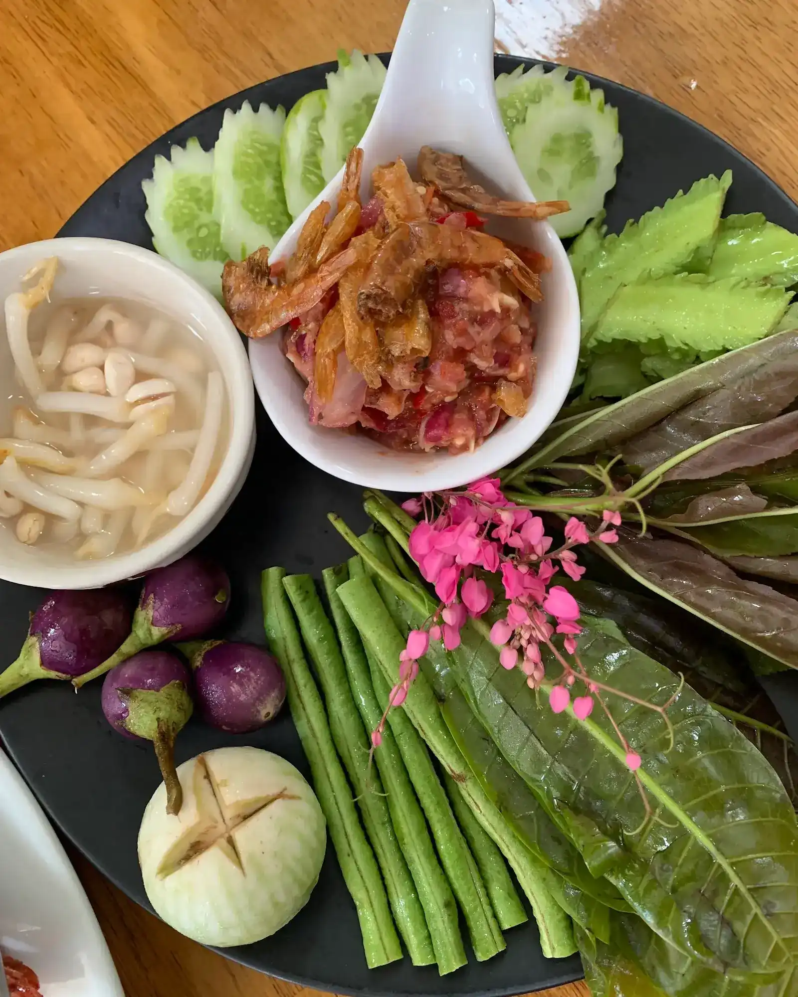 A chilli relish topped with crisp shrimp in a white spoon, on a black plate with cucumber, long beans, winged beans, Thai eggplants, fresh leaves, pink flowers and a bowl of bean sprouts
