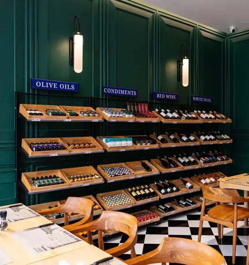 Dark green panelled grocer wall with navy signs over wooden shelves of olive oils and condiments, wooden chairs on a black and white floor