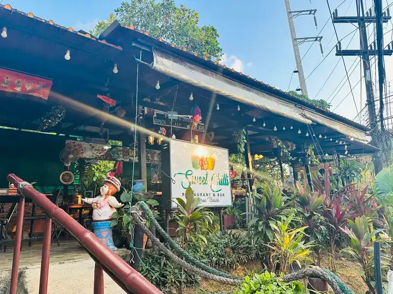 The front of Sweet Chilli: its sign, string lights and a garden of tropical plants | หน้าร้าน Sweet Chilli ป้ายร้าน ไฟประดับ และสวนไม้เขตร้อน