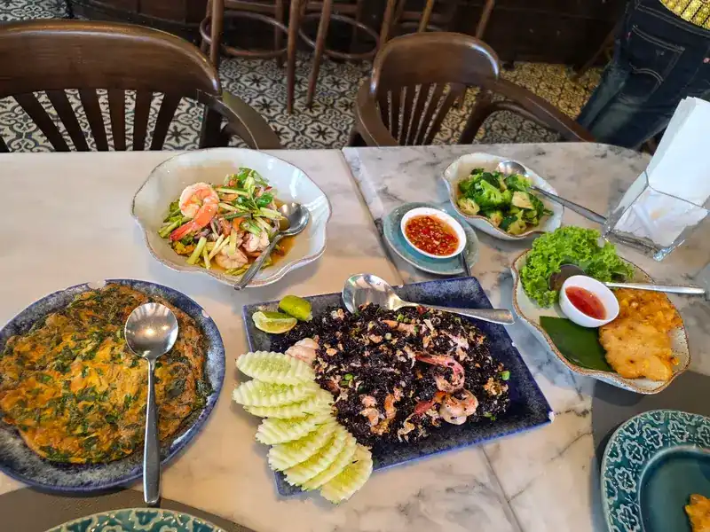 Dishes shared on a marble table: a shrimp salad, black fried rice with shrimp, an omelette, fried cakes and stir-fried greens