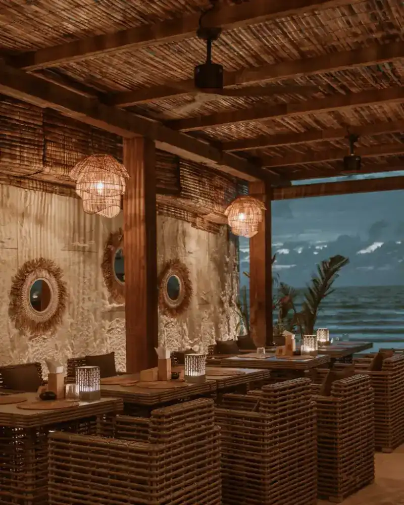 The restaurant at dusk: rattan chairs, woven lamps and round mirrors under a reed roof, open to the sea