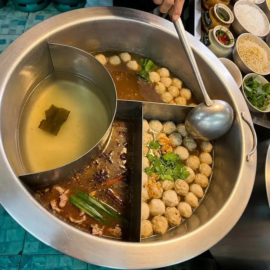 A round steel pot split into compartments: a clear broth, a dark spiced broth with star anise and pandan leaf, and meatballs ready for the ladle.