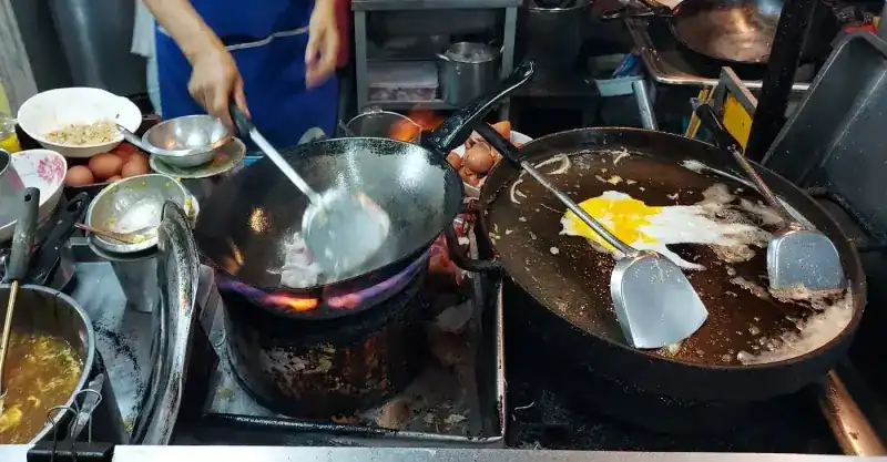 Two pans over the gas flame at the stall: one being tossed, one frying an egg