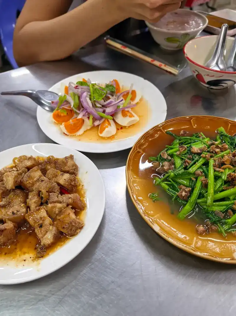 Three shared plates on a steel table: an egg salad with red onion, a pork dish and stir-fried morning glory
