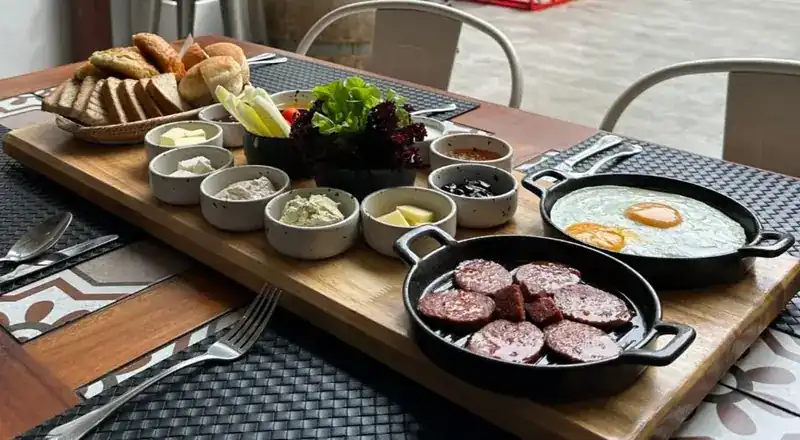 Turkish breakfast on a long wooden board: bread, small bowls of spreads, cheese and olives, fried eggs and a pan of sliced sucuk