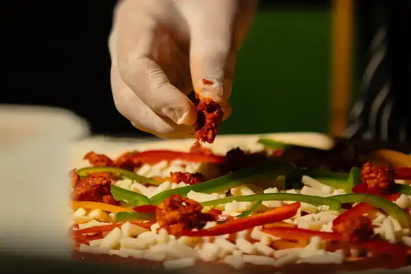 A gloved hand topping a pizza with peppers and cheese