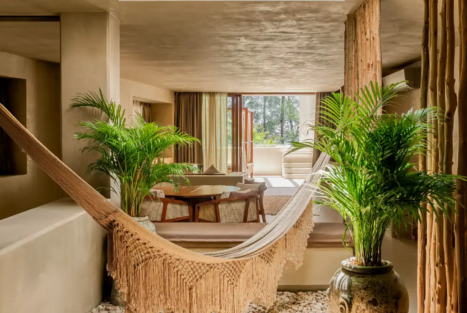 A woven hammock and palms in a sand-coloured room at Café del Mar Phuket, open to the trees outside