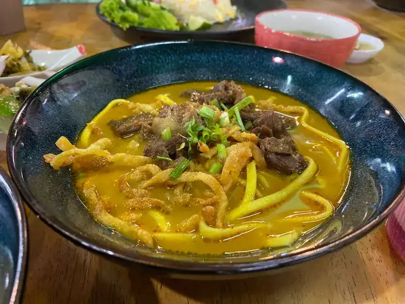 A dark bowl of khao soi: yellow curry broth, egg noodles, pieces of meat, spring onion and crisp noodles on top