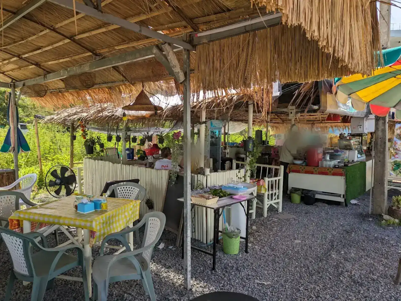 The open-air dining area at Art's Kitchen: a table with a yellow checked cloth on a gravel floor, a thatched roof, and the kitchen counter at the back