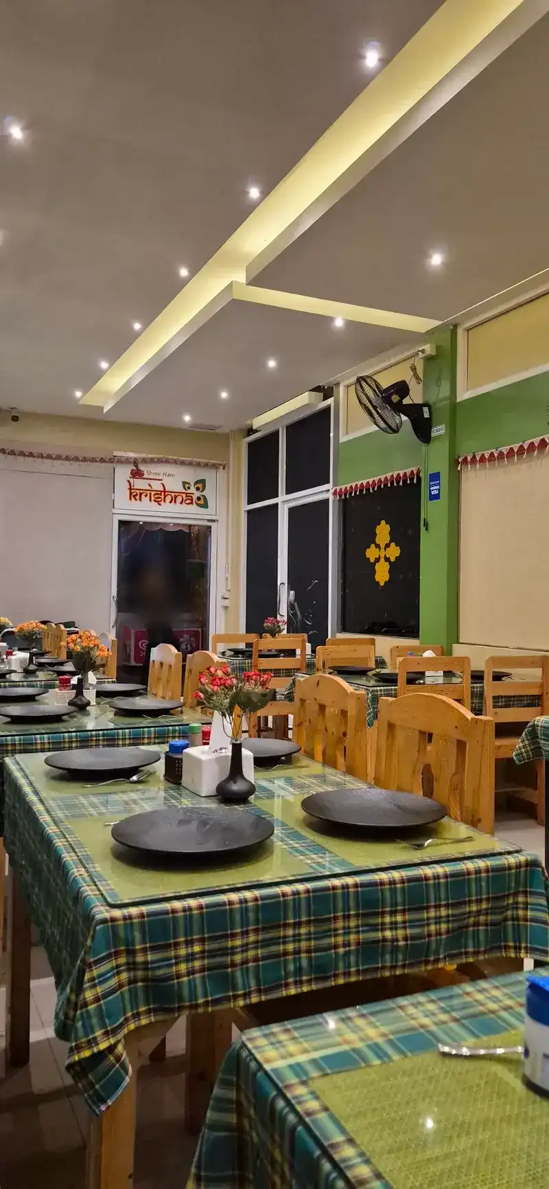 Rows of tables with green check cloths and wooden chairs, the Shree Hare Krishna sign over the door