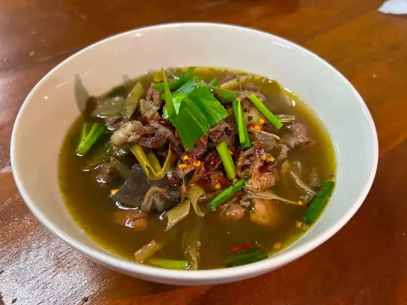 A bowl of herb broth with meat, spring onion, herbs and chilli flakes on a wooden table