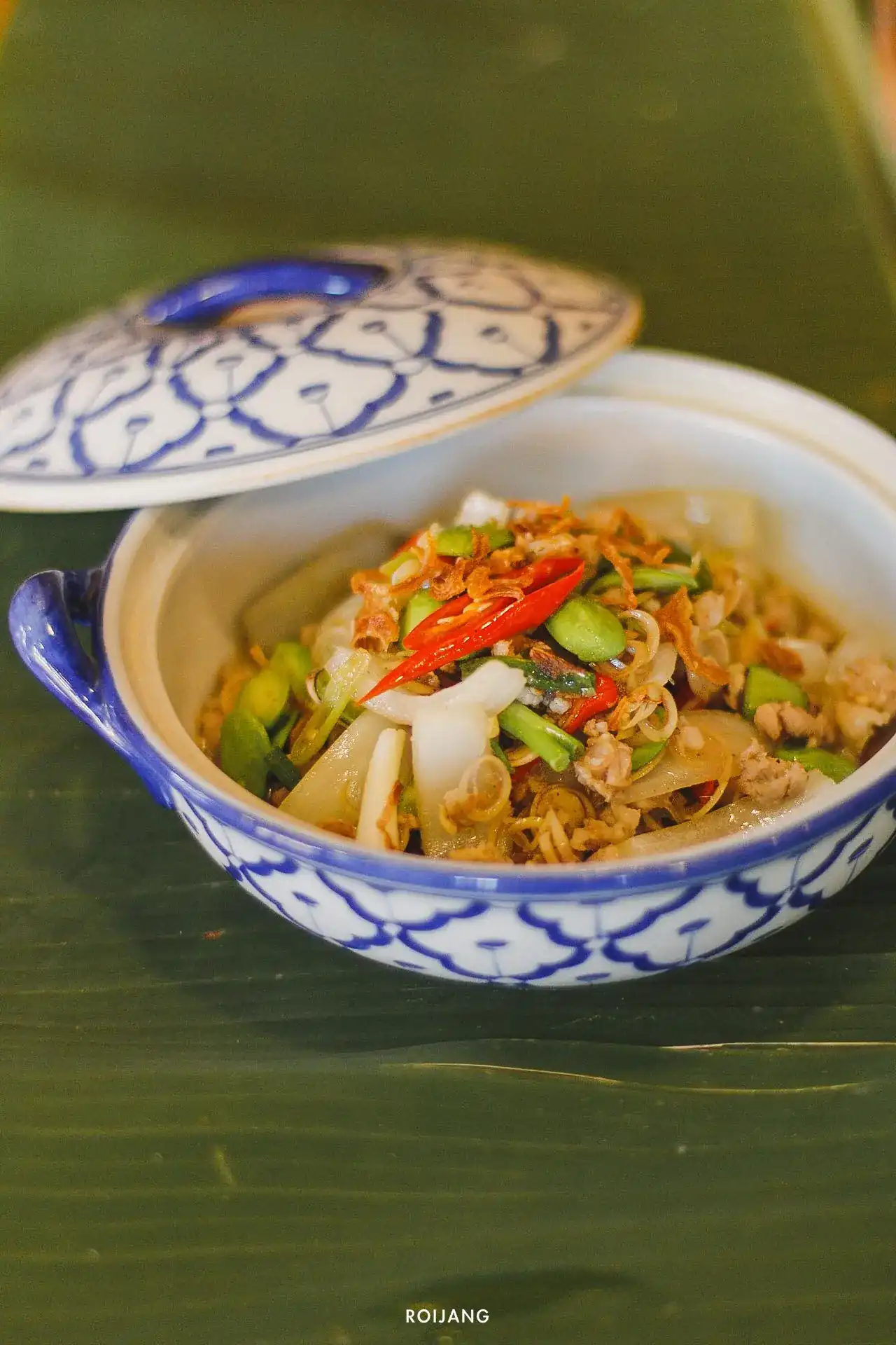 A stir-fry of green beans, minced pork, lemongrass and red chilli in a blue-and-white lidded bowl on a banana leaf