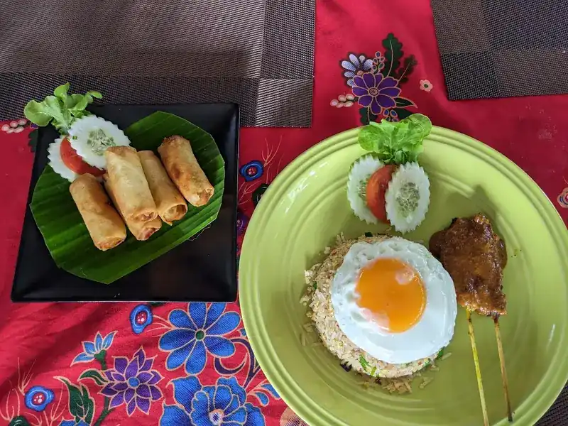 Spring rolls, and fried rice with a fried egg and satay skewers, on a red flowered tablecloth