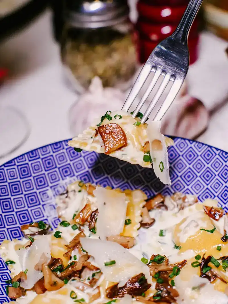 A fork lifting a filled pasta parcel with mushrooms and shaved cheese from a blue patterned bowl