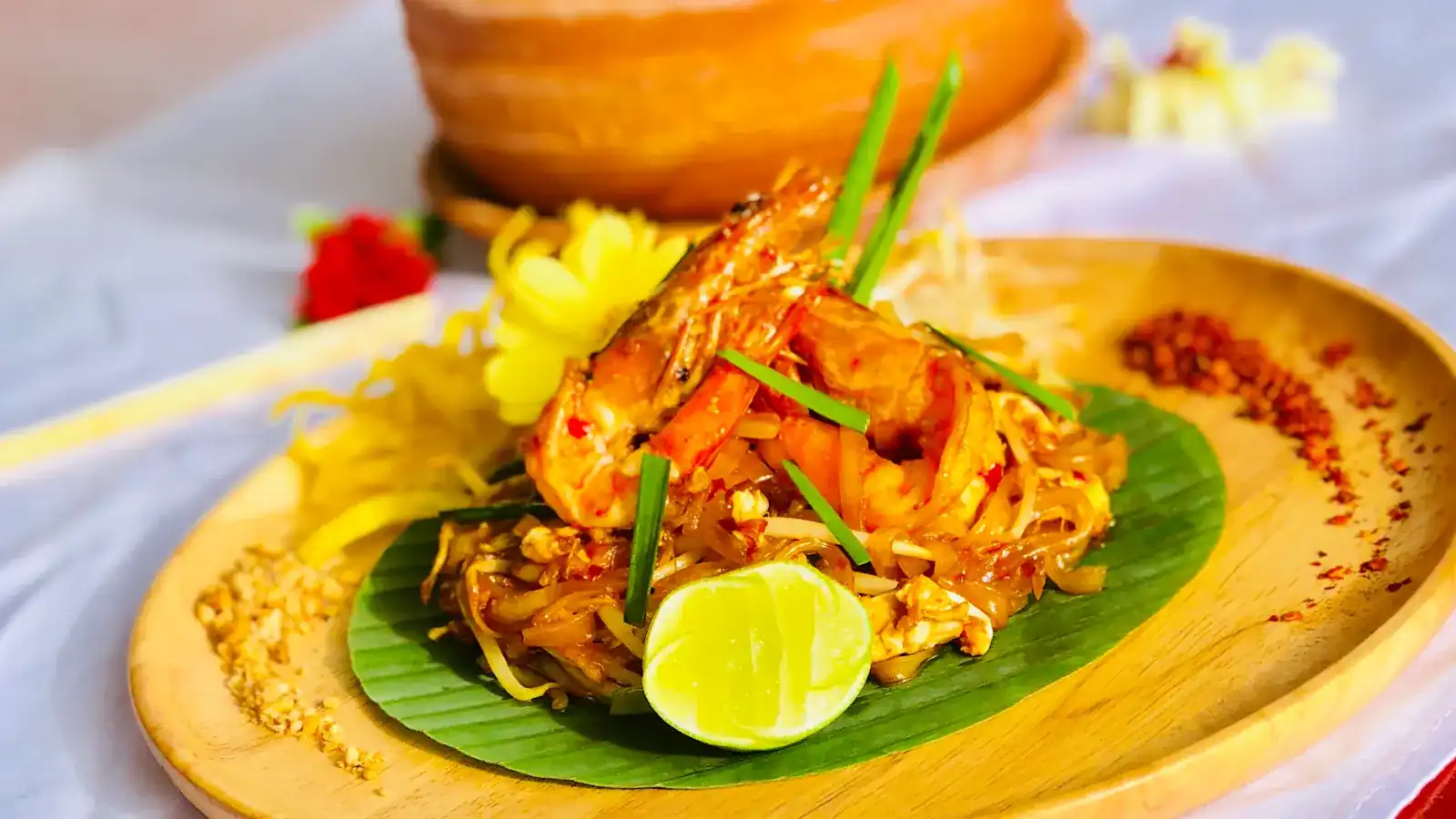 Pad thai topped with prawns on a banana leaf, with lime, crushed peanuts and chilli flakes on a wooden plate